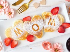 Mother's Day Gift: Heart shaped pancakes with MOM letters. Mothers Day breakfast concept. Top view table scene with a white wood background.