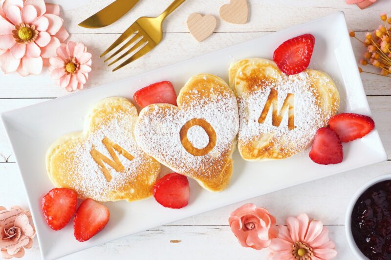 Mother's Day Gift: Heart shaped pancakes with MOM letters. Mothers Day breakfast concept. Top view table scene with a white wood background.