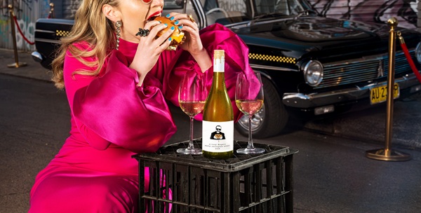 Woman Dinner: Woman in a pink outfit taking a bit of a burger with a bottle of white wine and two wine glasses in front of her