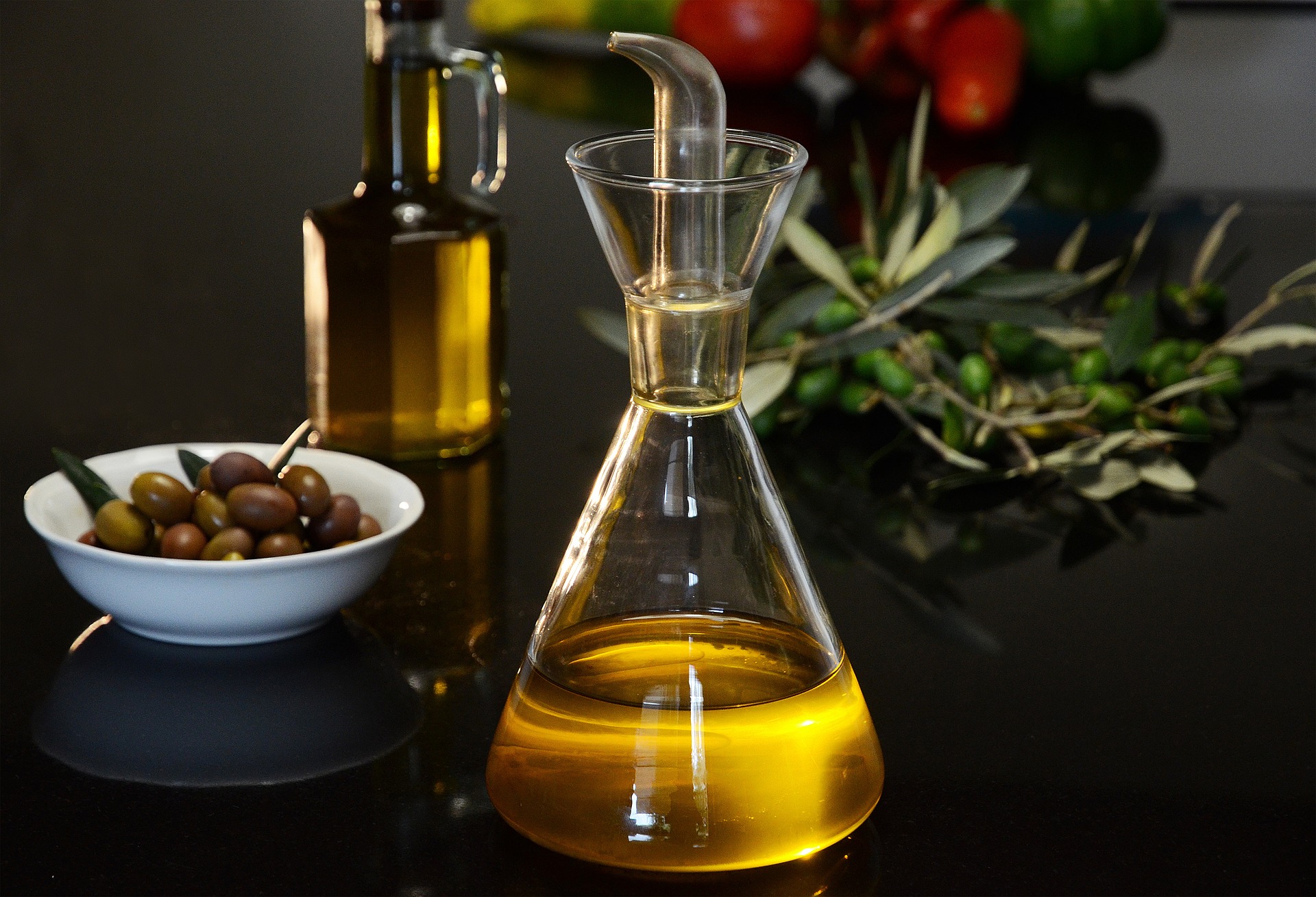 Eating Well: Olive oil bottle in the foreground with bowl of olives, rosemary and tomatoes in the background