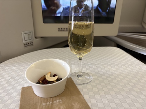 EVA Air Royal Laurel Class: Glass of white wine with small bowl of nuts in front of a screen on airplane