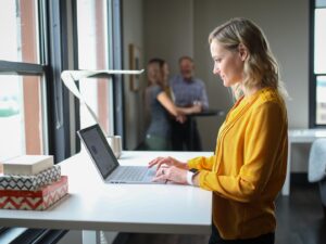 Progressive Desk: Woman at a standing desk on her laptop with two men in the background
