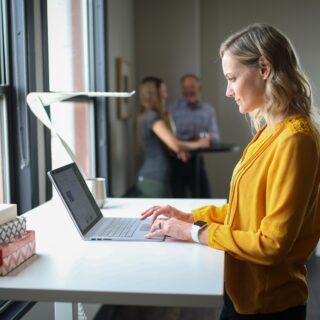 Progressive Desk: Woman at a standing desk on her laptop with two men in the background