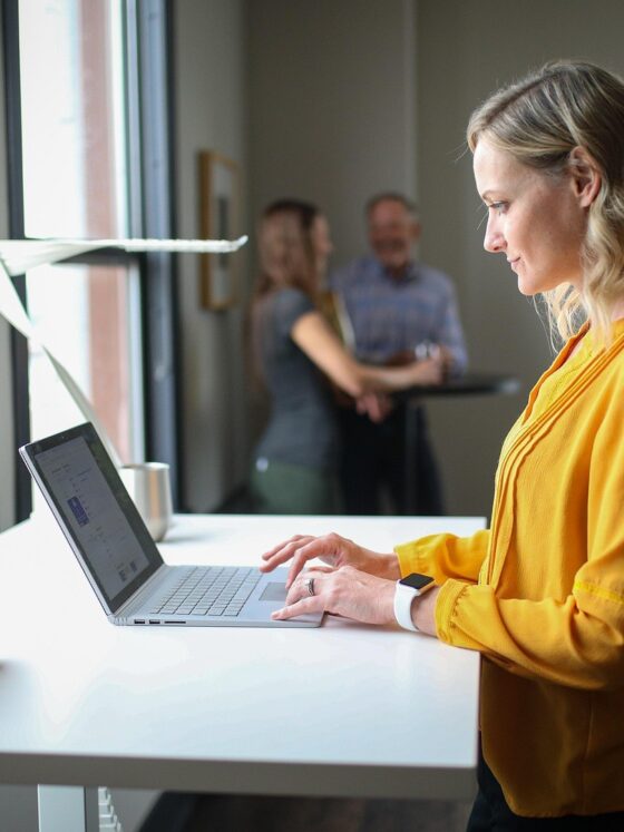 Progressive Desk: Woman at a standing desk on her laptop with two men in the background