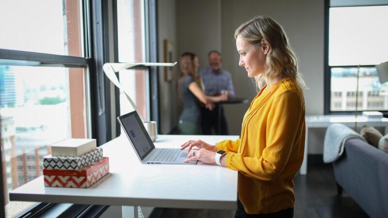 Progressive Desk: Woman at a standing desk on her laptop with two men in the background