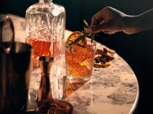 Whisky Tasting Party: woman's hand putting rosemary sprig into a glass of whisky - decanter nearby