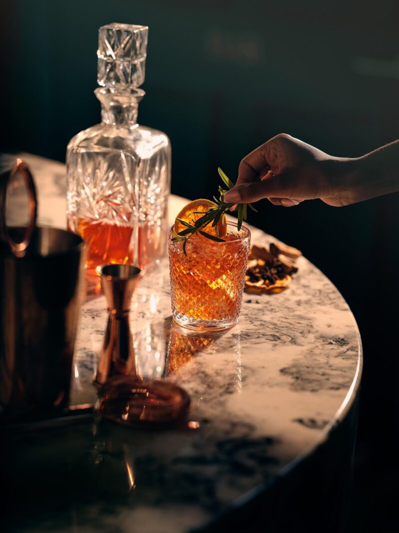 Whisky Tasting Party: woman's hand putting rosemary sprig into a glass of whisky - decanter nearby