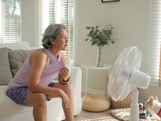 Hot Flash: Asian middle age old woman sit at sofa home feel hot flash fever use fan waving hand blowing wind face elder health care summer heat aging issue.