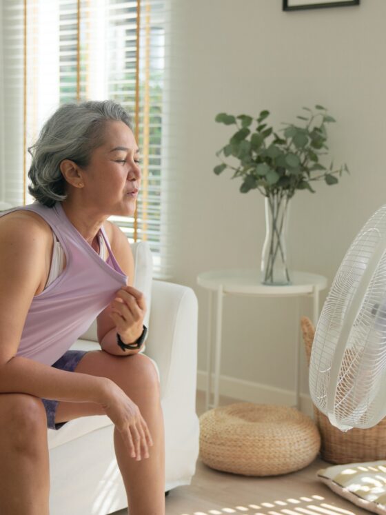 Hot Flash: Asian middle age old woman sit at sofa home feel hot flash fever use fan waving hand blowing wind face elder health care summer heat aging issue.