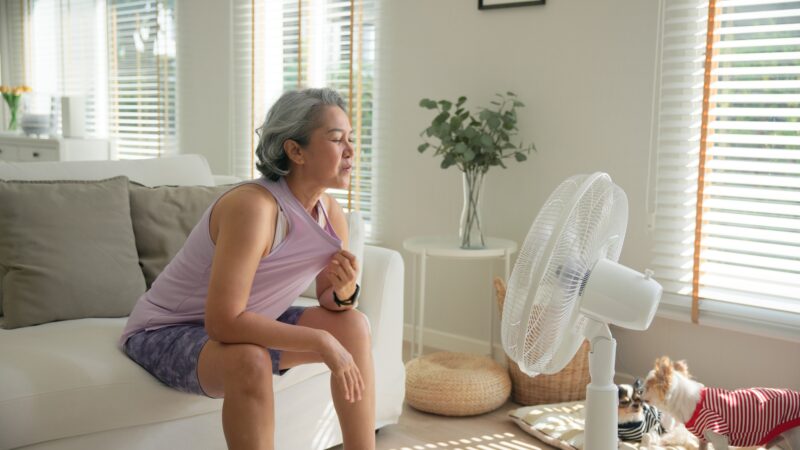 Hot Flash: Asian middle age old woman sit at sofa home feel hot flash fever use fan waving hand blowing wind face elder health care summer heat aging issue.