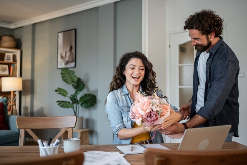 Busy Husband: adult man surprising his mid adult wife with a stunning bouquet of flowers while working from home, creating a heartwarming moment filled with love and appreciation