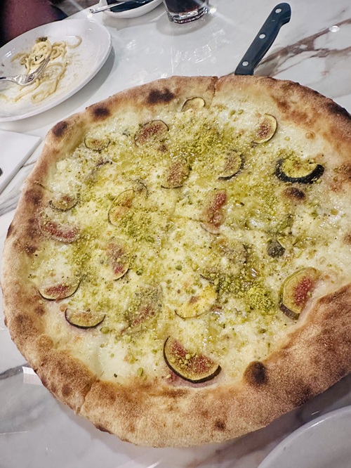 Vivo Avanti: Il Fugo, a white pizza with Gorgonzola cheese, figs, pistachio, and honey on a plate with an empty pasta plate in the background