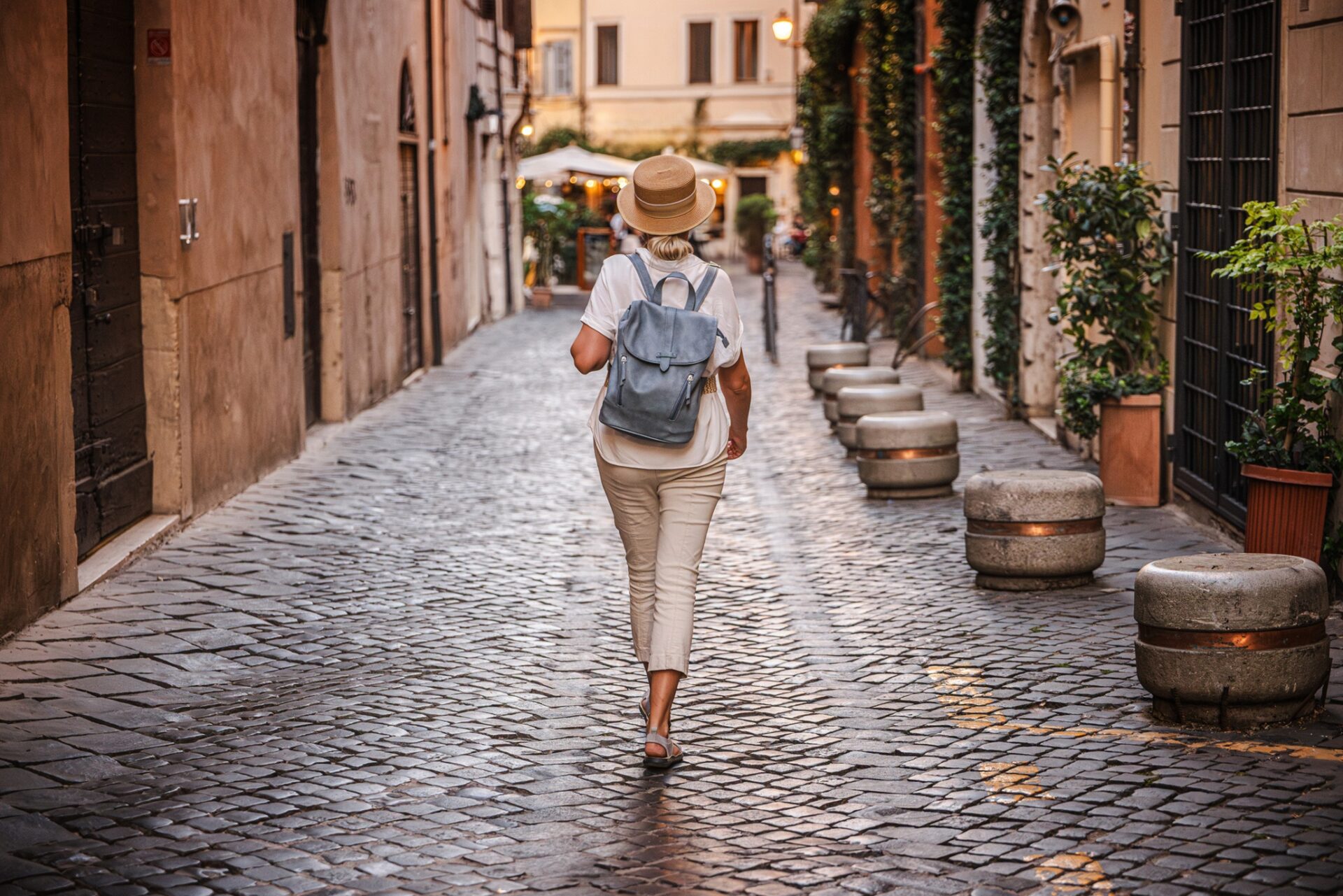 Women's Travel: Middle-aged woman wearing a straw hat and backpack walking on a cobblestone street in Rome with warm evening lights and historic atmosphere. High quality photo