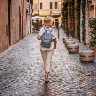 Women's Travel: Middle-aged woman wearing a straw hat and backpack walking on a cobblestone street in Rome with warm evening lights and historic atmosphere. High quality photo