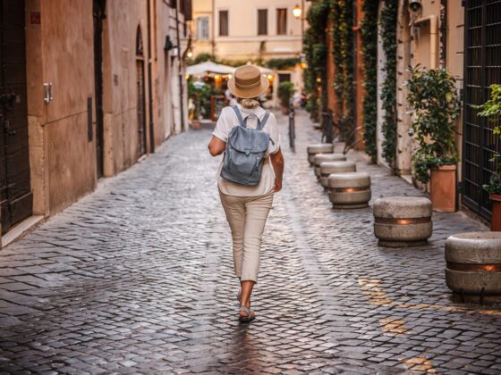 Women's Travel: Middle-aged woman wearing a straw hat and backpack walking on a cobblestone street in Rome with warm evening lights and historic atmosphere. High quality photo