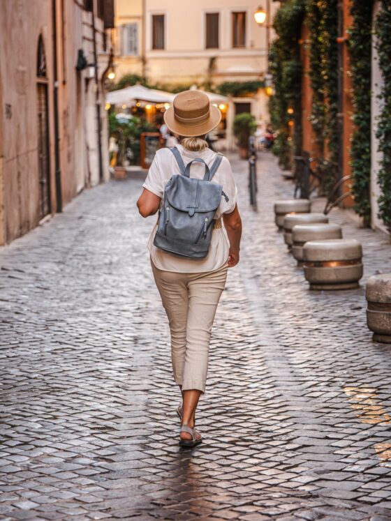 Women's Travel: Middle-aged woman wearing a straw hat and backpack walking on a cobblestone street in Rome with warm evening lights and historic atmosphere. High quality photo