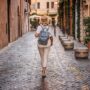 Women's Travel: Middle-aged woman wearing a straw hat and backpack walking on a cobblestone street in Rome with warm evening lights and historic atmosphere. High quality photo