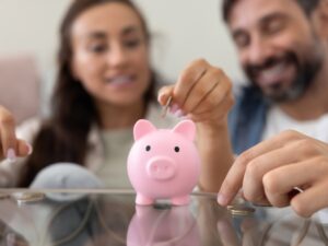 Smart Money Habits: Close up of hands throwing cash into pink piggybank with happy family couple in background. Husband and wife saving money for pension, making investment for future income, profit