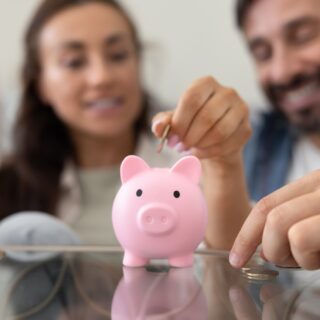 Smart Money Habits: Close up of hands throwing cash into pink piggybank with happy family couple in background. Husband and wife saving money for pension, making investment for future income, profit