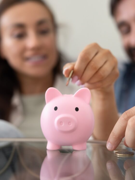 Smart Money Habits: Close up of hands throwing cash into pink piggybank with happy family couple in background. Husband and wife saving money for pension, making investment for future income, profit