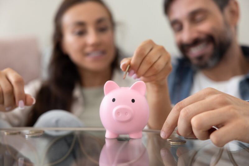 Smart Money Habits: Close up of hands throwing cash into pink piggybank with happy family couple in background. Husband and wife saving money for pension, making investment for future income, profit