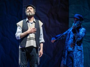 Fiddler On The Roof In Yiddish: Tevye singing with his hand over his heart with the fiddler in the background