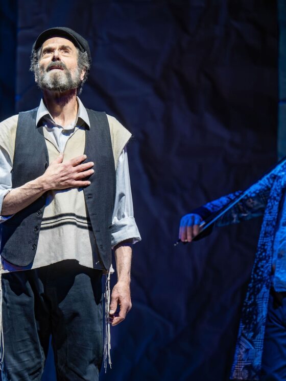 Fiddler On The Roof In Yiddish: Tevye singing with his hand over his heart with the fiddler in the background