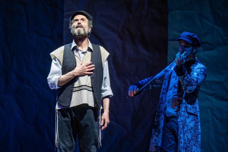 Fiddler On The Roof In Yiddish: Tevye singing with his hand over his heart with the fiddler in the background