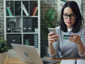 Pay Online: Woman in front of a laptop holding her mobile phone and bank card