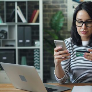Pay Online: Woman in front of a laptop holding her mobile phone and bank card