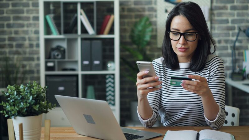 Pay Online: Woman in front of a laptop holding her mobile phone and bank card