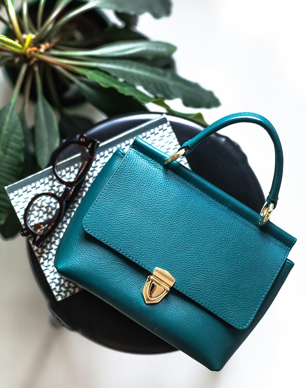 Timeles Handbags: Teal handbag on top of agenda with glasses beside it and plant.