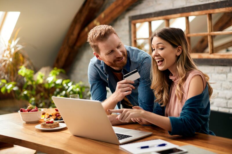 Credit Card: Happy woman pointing on something on laptop screen while shopping on the internet with her husband at home.