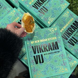 vikram vij: jmy new indian kitchen, cookbooks in a pile with a hand holding Gunpowder Prawns with Coconut Masala