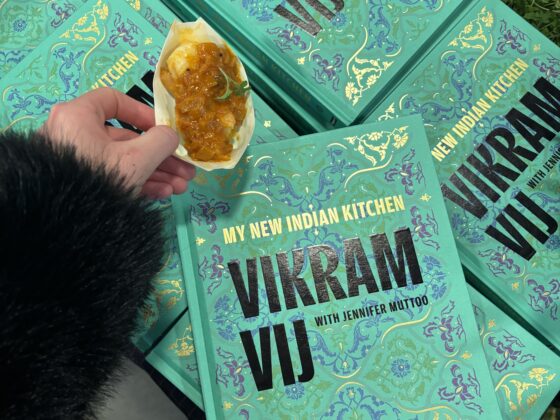 vikram vij: jmy new indian kitchen, cookbooks in a pile with a hand holding Gunpowder Prawns with Coconut Masala