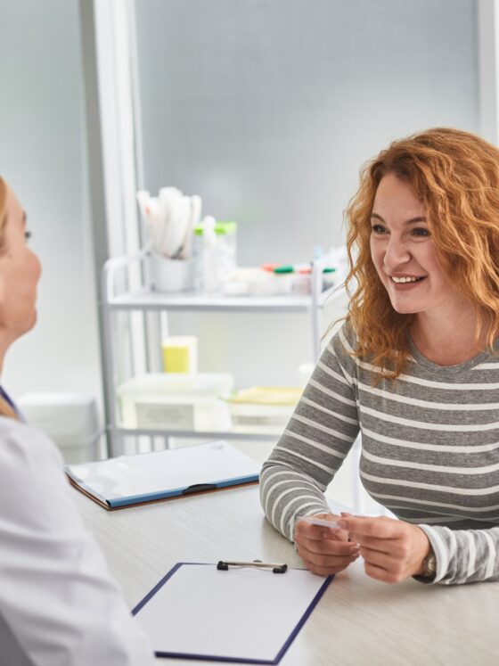 BHRT: Good news. Portrait of mature lady sitting at the table and having consultation with gynecologist. She is holding visiting card and smiling
