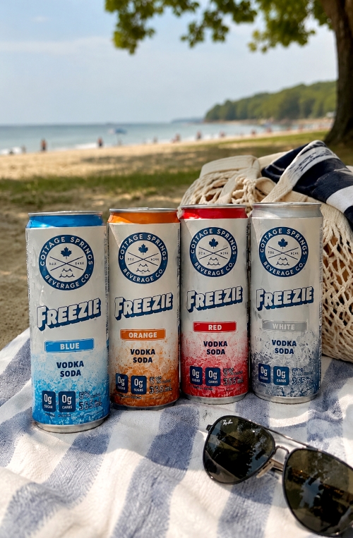 RTDs: Cottage Springs Vodka Soda Freezie cans on a beach blanket on beach next to sunglasses