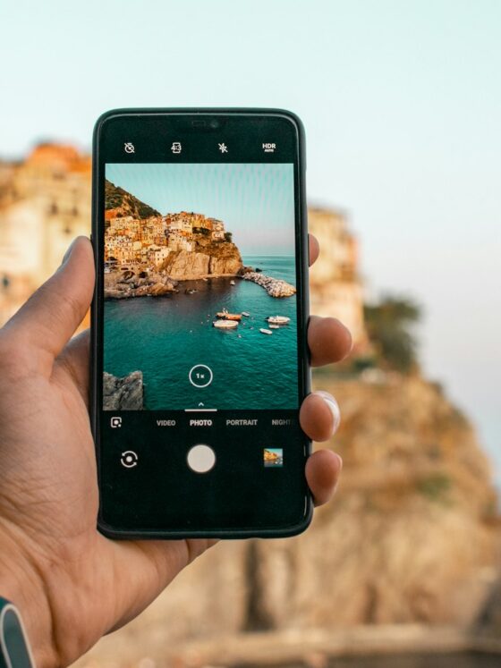 iPhone 18 Pro Max: Hand holding a smartphone to photograph a rocky coastline and the sea