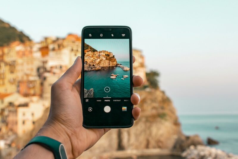 iPhone 18 Pro Max: Hand holding a smartphone to photograph a rocky coastline and the sea